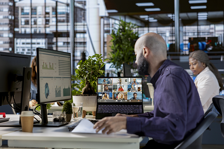 A man works at a desk with a laptop showing a video conference, alongside a desktop monitor displaying charts in a modern open-plan office with other people working in the background.