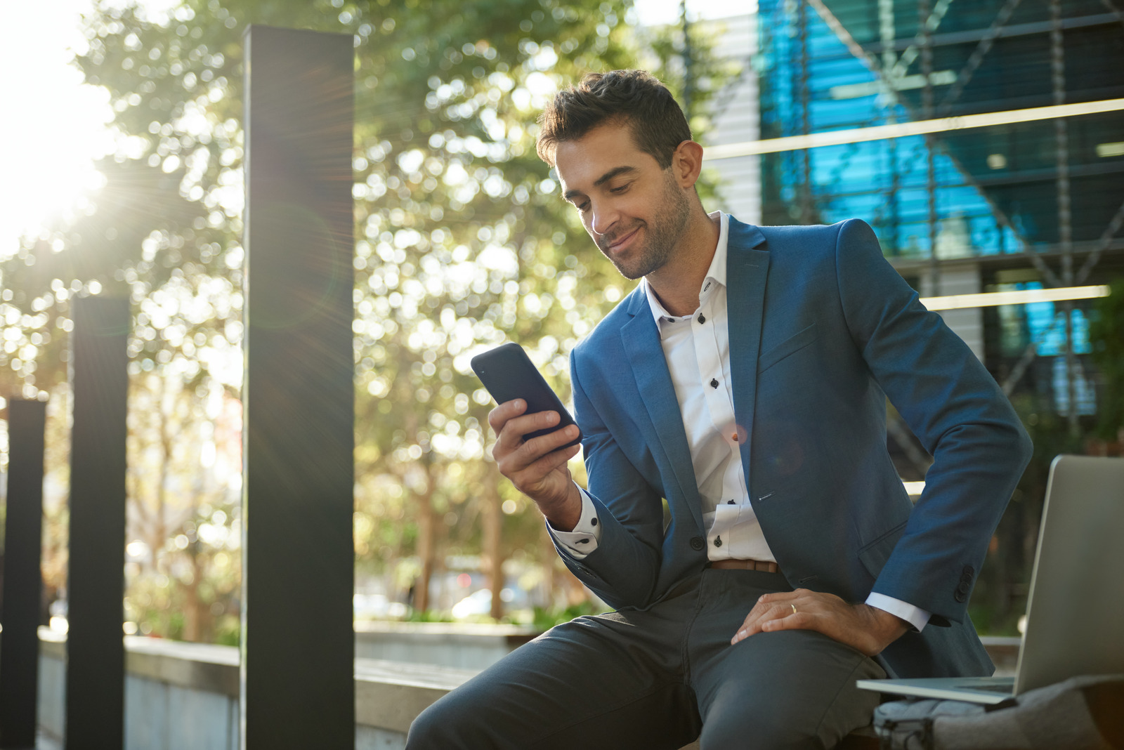A man in a blue blazer sits outdoors, smiling while looking at his phone—perhaps discovering the UCaaS Solutions Benefits—in a modern urban setting with sunlight filtering through trees and a glass building in the background.