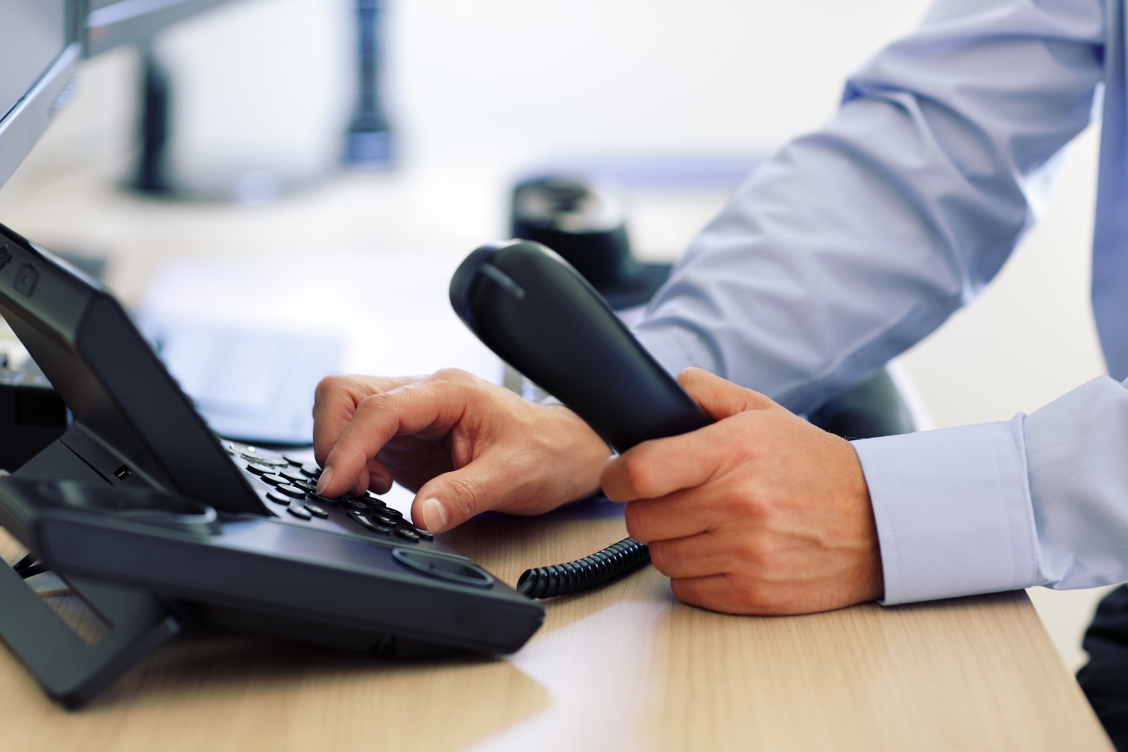 A person in a blue shirt sits at a desk, holding a phone handset in one hand and dialing numbers on the office phone with the other, exploring POTS replacement benefits for their communication needs.