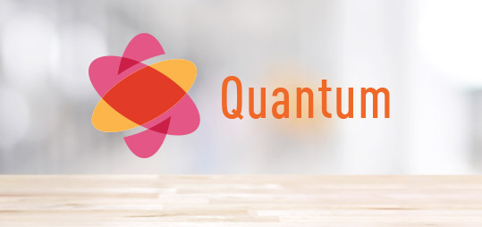 A stylized atom logo in pink, orange, and red appears next to the word Quantum in bold orange text, with STACKedge subtly displayed on a blurred light background above a wooden surface.
