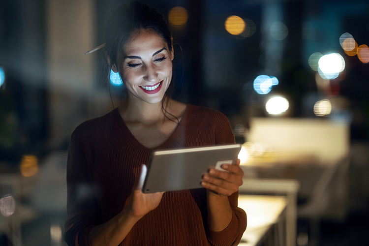 A woman smiles while using a tablet at night in a softly lit room. She has a pencil behind her ear and wears a casual sweater, with blurred city lights visible in the background.